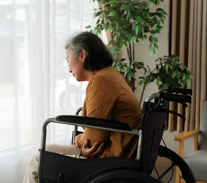 Senior woman leaning forward while seated in a wheelchair near a home window, depicting daily mobility challenges, aging, accessibility needs, elderly support, caregiving and residential healthcare.