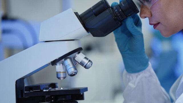 Close up of a scientist looking through microscope eyepiece with objective lens in focus, wearing glove and safety glasses in a research laboratory