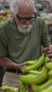 An elderly man browses bananas in a grocery store, carefully selecting ripe ones among the bunch.