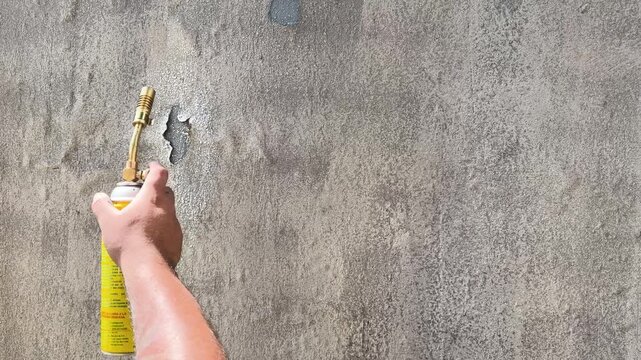 Hand uses blowtorch to create rough textures on gray plaster wall during surface work, lifts and spreads flame to cause paint to bubble