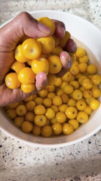 Person scoops yellow nance fruits from bowl of water on speckled kitchen counter