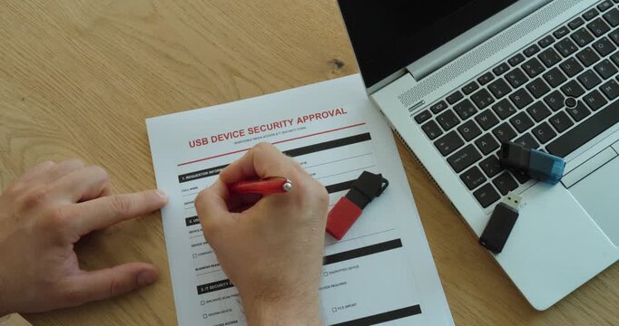 Person completing a USB device security approval form beside a laptop and flash drives, representing cybersecurity, data protection and workplace access control.