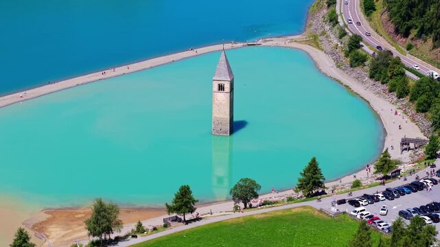 Iconic aerial view of the Curon bell tower rising from Lake Resia, with green alpine fields, mountain slopes and turquoise reservoir water in northern Italy