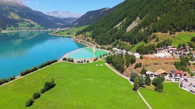 Scenic drone shot of the famous submerged church tower at Lake Resia, set against bright turquoise water and the mountain landscape of South Tyrol, Italy