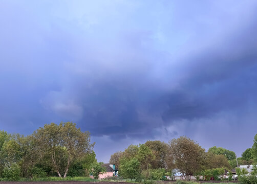 A dramatic sky with dark clouds looms over a rural landscape featuring lush green trees and small houses, suggesting an impending storm.