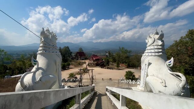Twin white lion statues flank the stairway to a hilltop temple overlooking a broad valley. Mountain ridges stretch across the horizon beneath a partly cloudy sky in Pai Thailand