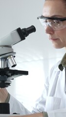 Female scientist in a lab coat and protective glasses examining samples through a microscope in a...