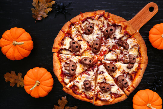 Halloween skull pizza. Top view table scene on a black wood background. Pepperoni and cheese with spooky mushroom skulls.