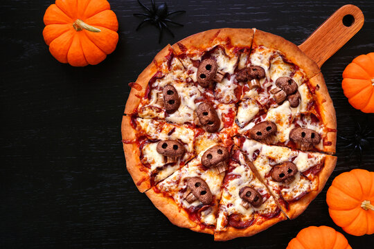 Halloween skull pizza. Overhead view table scene on a black wood background. Pepperoni and cheese with spooky mushroom skulls. Copy space.