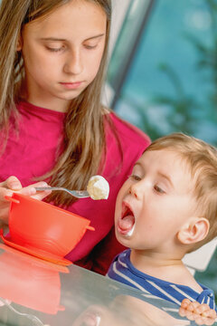 Older sister feeding her little brother with a spoon at home.