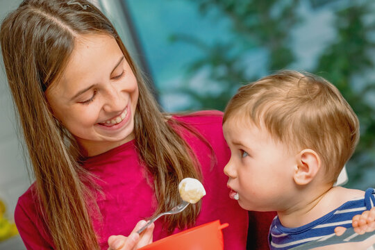 Older sister feeding her little brother with a spoon at home.
