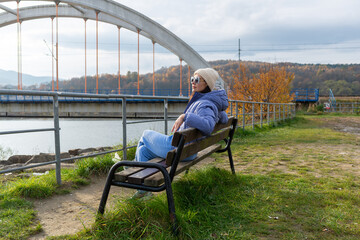Female traveler wearing a blue puffer jacket, beige knitted beanie and sunglasses sits peacefully on a bench overlooking Jezioro Mucharskie. Repeating concrete arches, autumn woodland and water form a © Volha