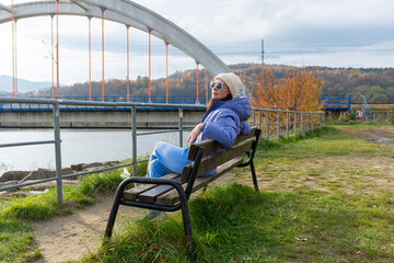 Adult woman in a blue puffer jacket, knit beanie and sunglasses relaxes on a wooden bench beside Jezioro Mucharskie in southern Poland. The large arched bridge, autumn hills and calm reservoir create  © Volha
