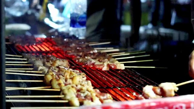 Thai street food vendor arranges pork skewers over glowing charcoal at a night market, showing authentic local cuisine, hands on food service, and small business takeaway culture.