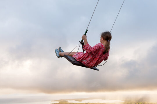 A little girl in a pink dress swings high above a misty landscape at sunset, conveying childhood freedom, joy and adventure against dramatic clouds and soft golden light