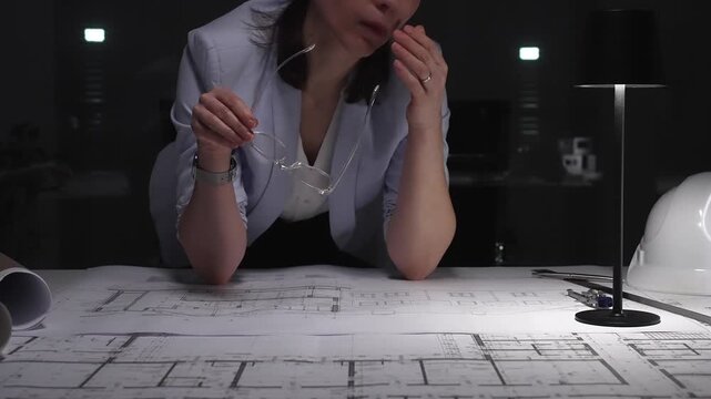 Tired woman architect feeling stress and fatigue while working on project blueprints at her desk in a dark office at night. Business and architect people concept