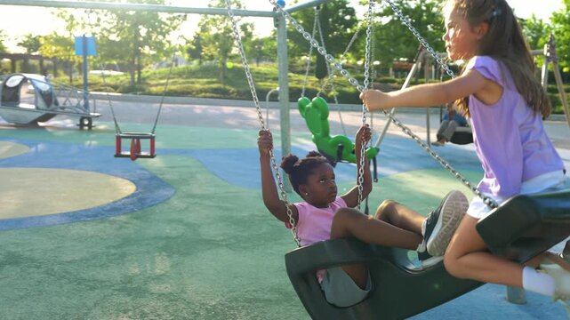 Two cheerful multiethnic children playing on a swing set. An african american and a caucasian girl having fun together outdoors in a park