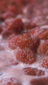 A large vessel contains boiling strawberries, emitting steam and bubbles. The thickening mixture is stirred with a spoon to ensure the strawberries cook evenly and turn into jam