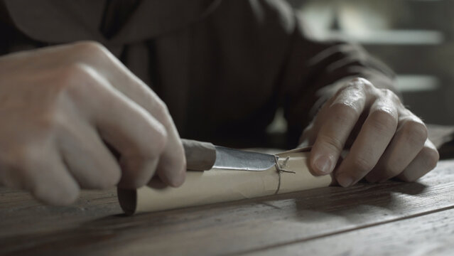 Medieval monk hands cutting a cord knot on a rolled parchment with a penknife on a wooden table inside a scriptorium