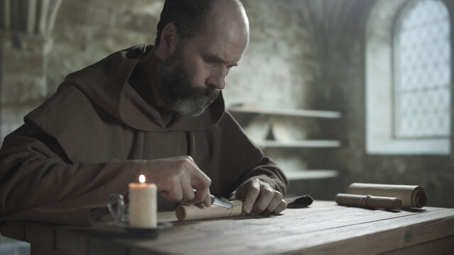 Medieval monk cutting a twine string on a parchment paper scroll with a knife at a wooden table near a burning candle inside a scriptorium