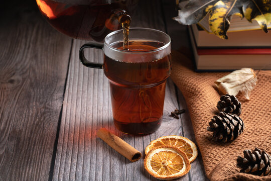 Hot tea pouring into a glass mug with orange and cinnamon. Cozy autumn rustic vibe with books, pinecones and warm light