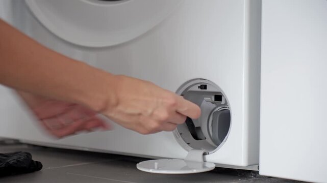 Woman removes cap from drain hose inside washing machine filter compartment and water flows out. Routine washer maintenance before cleaning drain filter