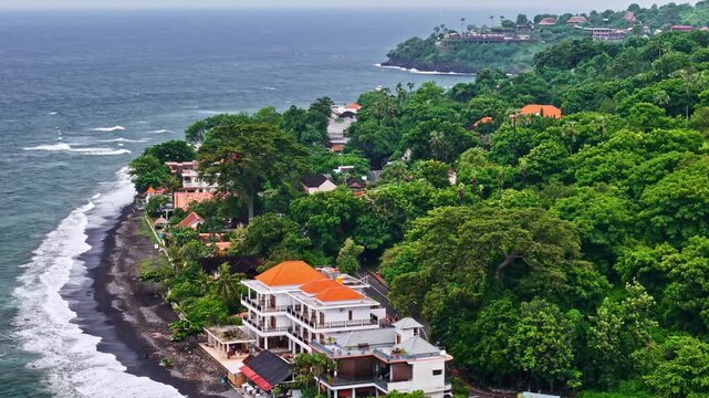 Aerial view of Amed Beach with black sand, lush tropical vegetation, and coastal villas with orange roofs in Bali, Indonesia.