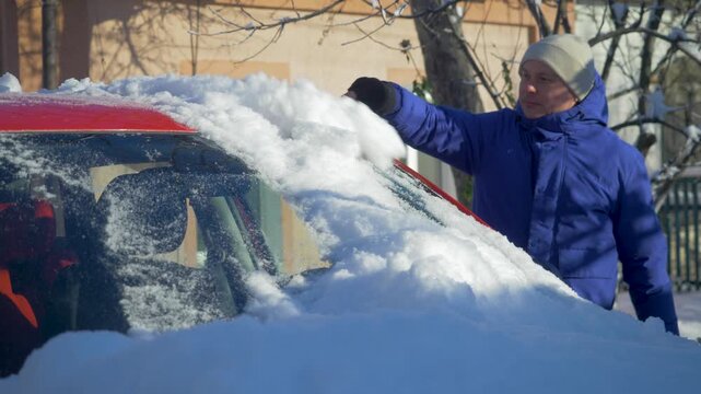 Man uses snow brush to clear front glass of red car covered by heavy winter precipitation. Frost and deep snow limit visibility. Severe cold creates demanding conditions for morning travel