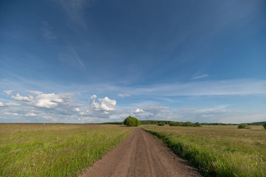 Dirt country road flanked by green fields under a wide blue sky