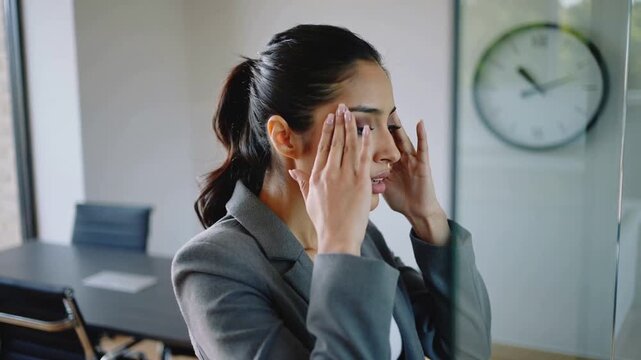 Standing woman in gray blazer rubbing temples at glass partition amid meeting pause
