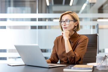 Businesswoman wearing glasses sitting at an office desk with a laptop, looking away and...