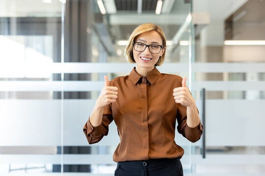 Smiling business woman wearing glasses and a brown shirt stands in a modern office, showing a double thumbs up gesture, expressing success, approval, and positivity