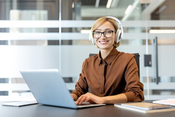 Fototapeta premium Smiling businesswoman wearing headphones and eyeglasses sitting at her desk in a modern office. Actively typing on a laptop. And looking at the camera. Conveying professionalism. Focus. And connection