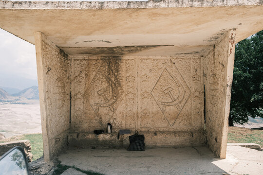 View of a weathered concrete bus stop featuring Soviet-era hammer and sickle reliefs on the back wall, with a thermos and a bag resting on the bench in Tajikistan.