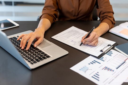 Woman's hands typing on laptop keyboard and writing on paper document with a pen, managing business paperwork and financial data in a modern office environment