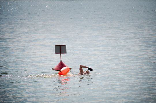 Open water swimmer passing navigation buoy with safety float, performing freestyle stroke in lake during endurance training. Concept of triathlon, outdoor sport and distance swimming
