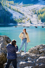 Photographer captures stylish young woman posing on rocks beside Lago di Braies, with rowing boats and autumn forest behind. Suitable for travel campaigns, tourism advertising, content creation themes © anatoliy_gleb