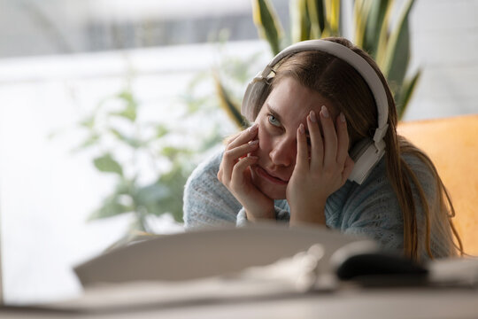 Stressed young Caucasian woman wearing headphones, covering her face with her hands in frustration while working from home, looking anxious and overwhelmed