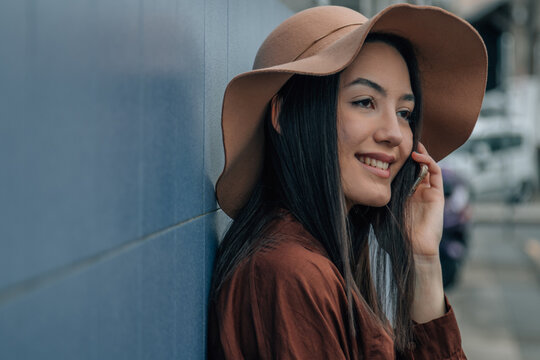 Smiling young Asian woman leaning against a wall while talking on smartphone, happy candid outdoor portrait