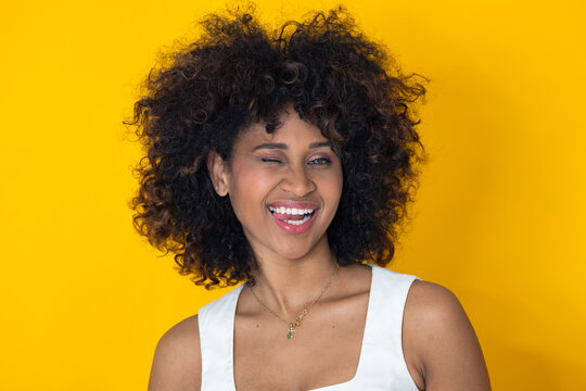 Portrait of a joyful mixed-race young woman winking and laughing against a vibrant yellow background, expressing happiness and confidence