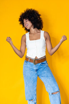 happy young latin woman celebrating success with fists raised, joyful expression and eyes closed, isolated on yellow background