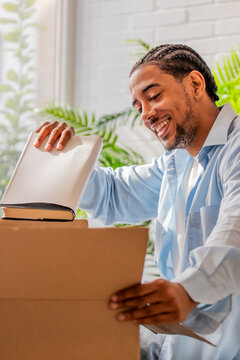 smiling young black man unpacking a book from a cardboard box while moving into his new home, surrounded by houseplants and natural light