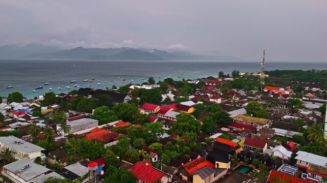 Aerial view of Gili Trawangan coastal town with red-roofed houses, lush greenery, and boats anchored in the bay near Pemenang, West Nusa Tenggara, Indonesia.