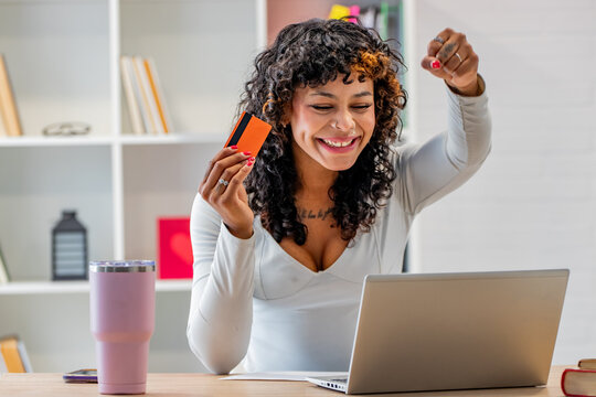 excited mixed-race woman celebrating the success of her online purchases with credit card in hand while looking at her laptop and cheering with her fist raised in her home office