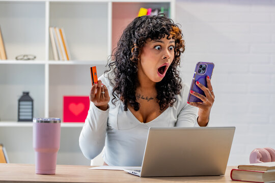 shocked mixed race woman with wide eyes and open mouth holding a credit card and smartphone, expressing surprise and disbelief while looking at her phone screen
