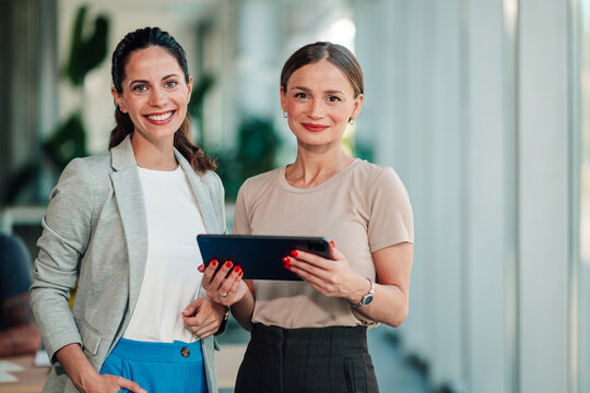 Two smiling women business professionals standing in modern office, collaborating with digital tablet