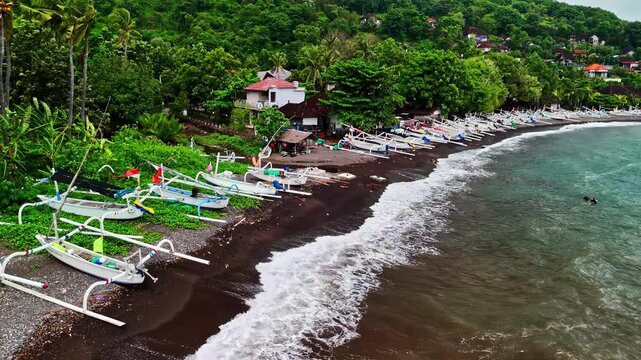 Aerial view of Amed Beach featuring numerous traditional outrigger boats lined up on the black sand shore surrounded by lush tropical greenery in Bali, Indonesia.