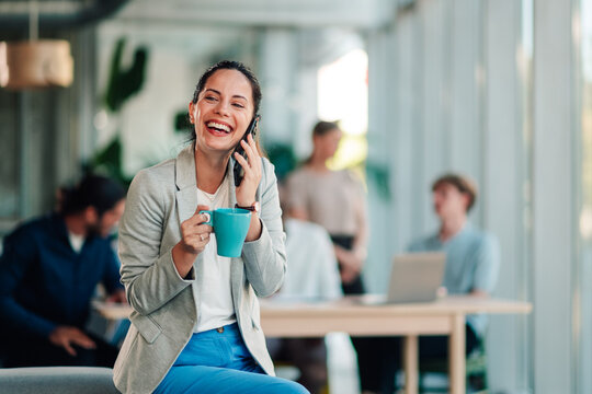 Professional woman laughing and talking on phone, holding coffee cup in a modern office working environment