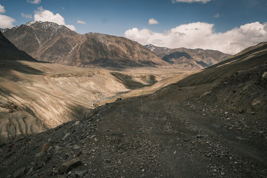 View of the rugged Bartang Valley with a winding dirt road in the foreground, surrounded by towering mountains under a partly cloudy sky in Chah Ab, Gorno-Badakhshan Autonomous Region, Tajikistan.
