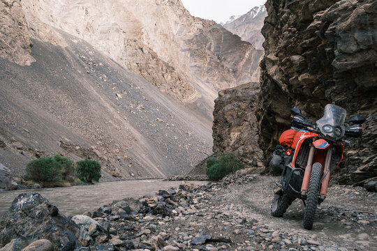 View of a motorcycle parked on a rocky trail beside the Bartang River, set against steep, rugged mountain slopes in Chah Ab, Gorno-Badakhshan Auto, Tajikistan.